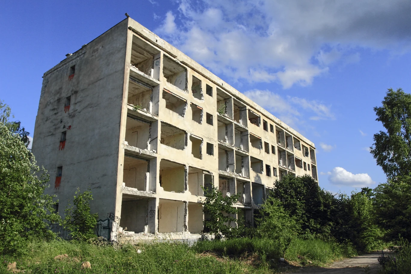 Zniszczony, pięciokondygnacyjny budynek mieszkalny o pustych wnętrzach i wypadających oknach, otoczony roślinnością i zaroślami, na tle niebieskiego nieba z chmurami.
