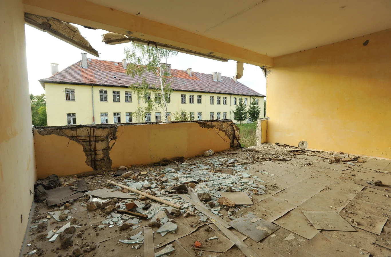 Zniszczone wnętrze budynku z dużym otworem w ścianie, gruzem oraz rozbitymi szybami na podłodze, przez wyłom widoczne żółte budynki z czerwoną dachówką i drzewa na tle spokojnej ulicy.