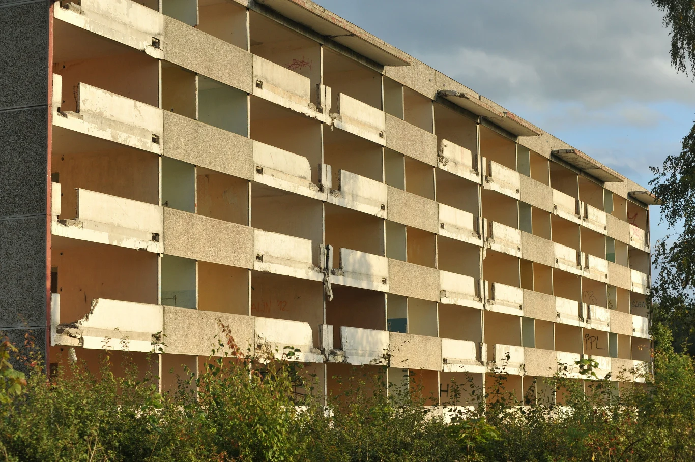 Opuszczony budynek mieszkalny z wieloma balkonami i brakiem szyb w oknach, oznaki zaniedbania i rozpadu, zarośnięte krzewy na pierwszym planie.