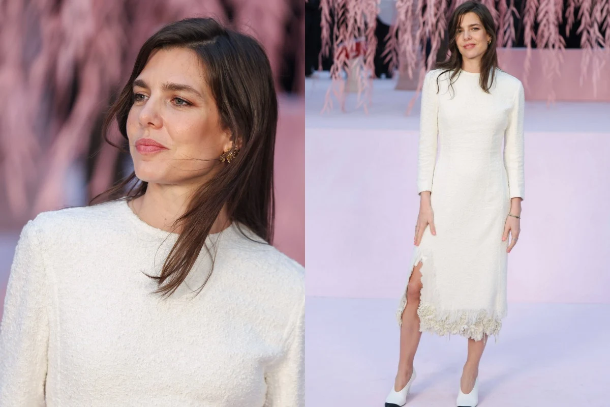 Kobieta w białej, eleganckiej sukience, stojąca na jasnym tle z różowymi dekoracjami, uśmiecha się subtelnie patrząc w bok. Sukienka sięga do kolan, z delikatnym rozcięciem na dole i koronką. Jasne buty na obcasie dopełniają stylizacji.