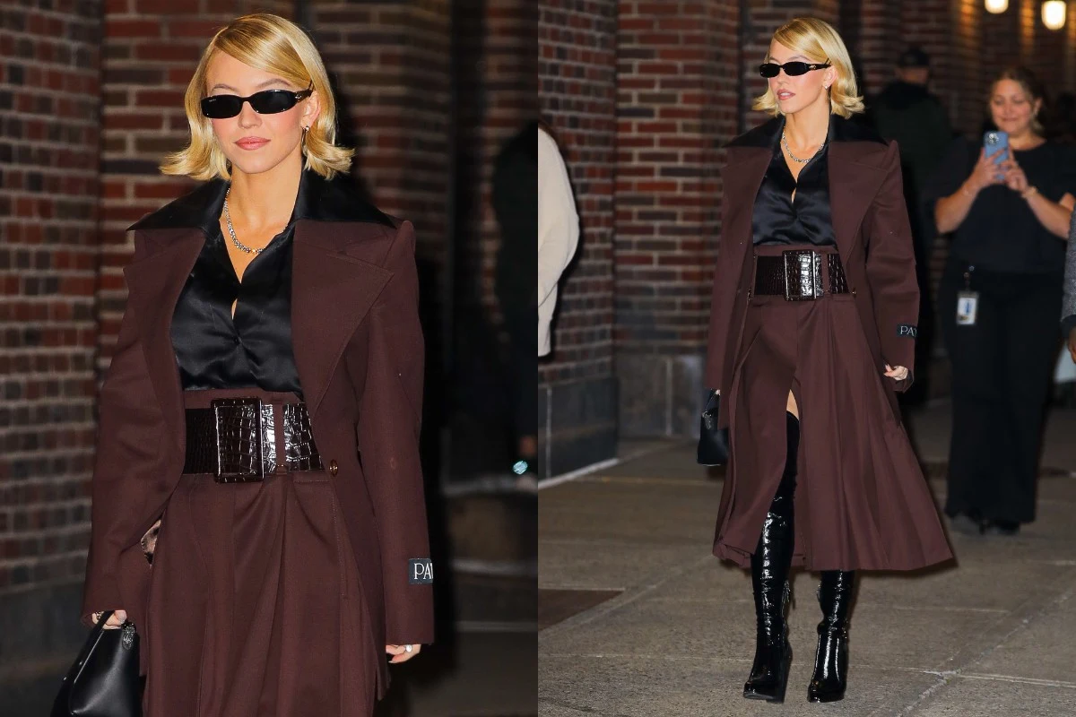 Stylowa kobieta w ciemnobrązowym płaszczu i połyskującej czarnej koszuli, z szerokim paskiem w talii oraz długimi, lakierowanymi kozakami. Ubrana w elegancki outfit z czarnymi okularami przeciwsłonecznymi, trzyma w ręce torebkę i stoi na chodniku przy ...