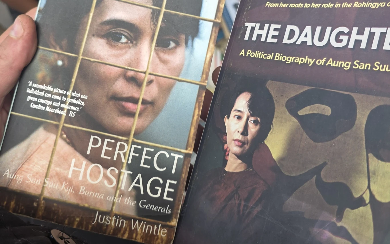 Dwie książki trzymane w rękach, obie poświęcone Aung San Suu Kyi, z jej portretami na okładkach, pierwsza z nich zatytułowana 'Perfect Hostage', druga 'The Daughter', widoczna kratka symbolizująca więzienie.