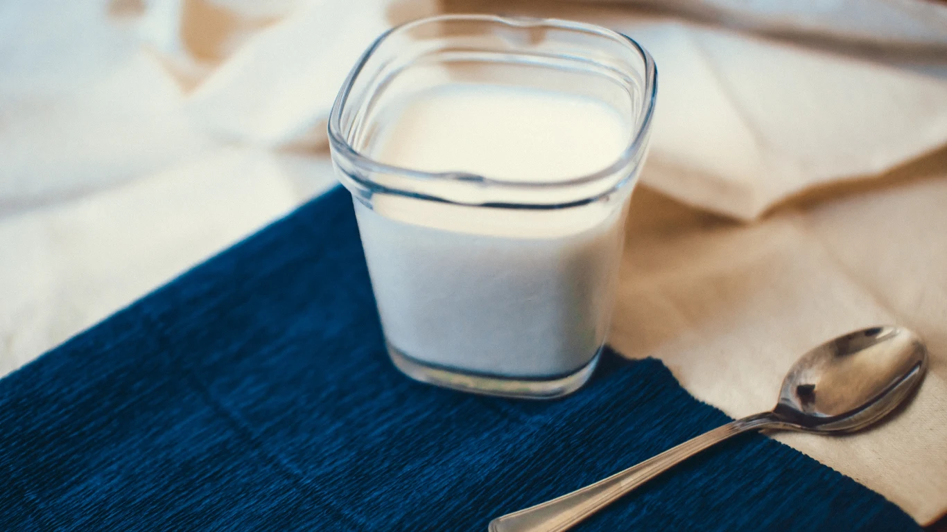 Eko jogurt naturalny Lublanka może zawierać fragmenty szkła / zdjęcie ilustracyjne Szklanka z białym jogurtem ustawiona na niebieskiej serwetce obok metalowej łyżeczki, tło w odcieniach beżu.