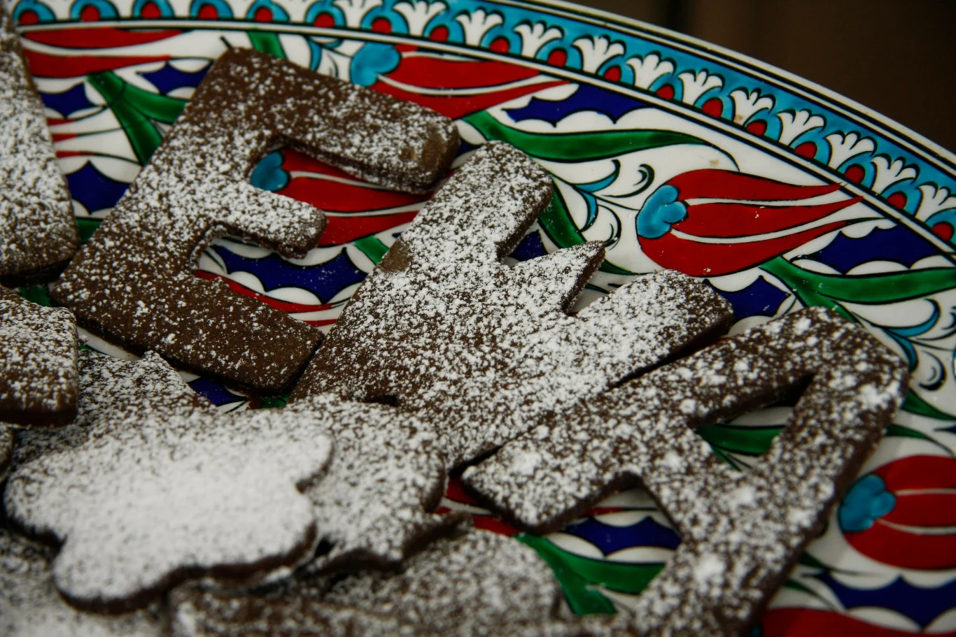 Czekoladowe ciasteczka w kształcie liter posypane cukrem pudrem, ułożone na dekoracyjnym talerzu z kolorowym, orientalnym wzorem.