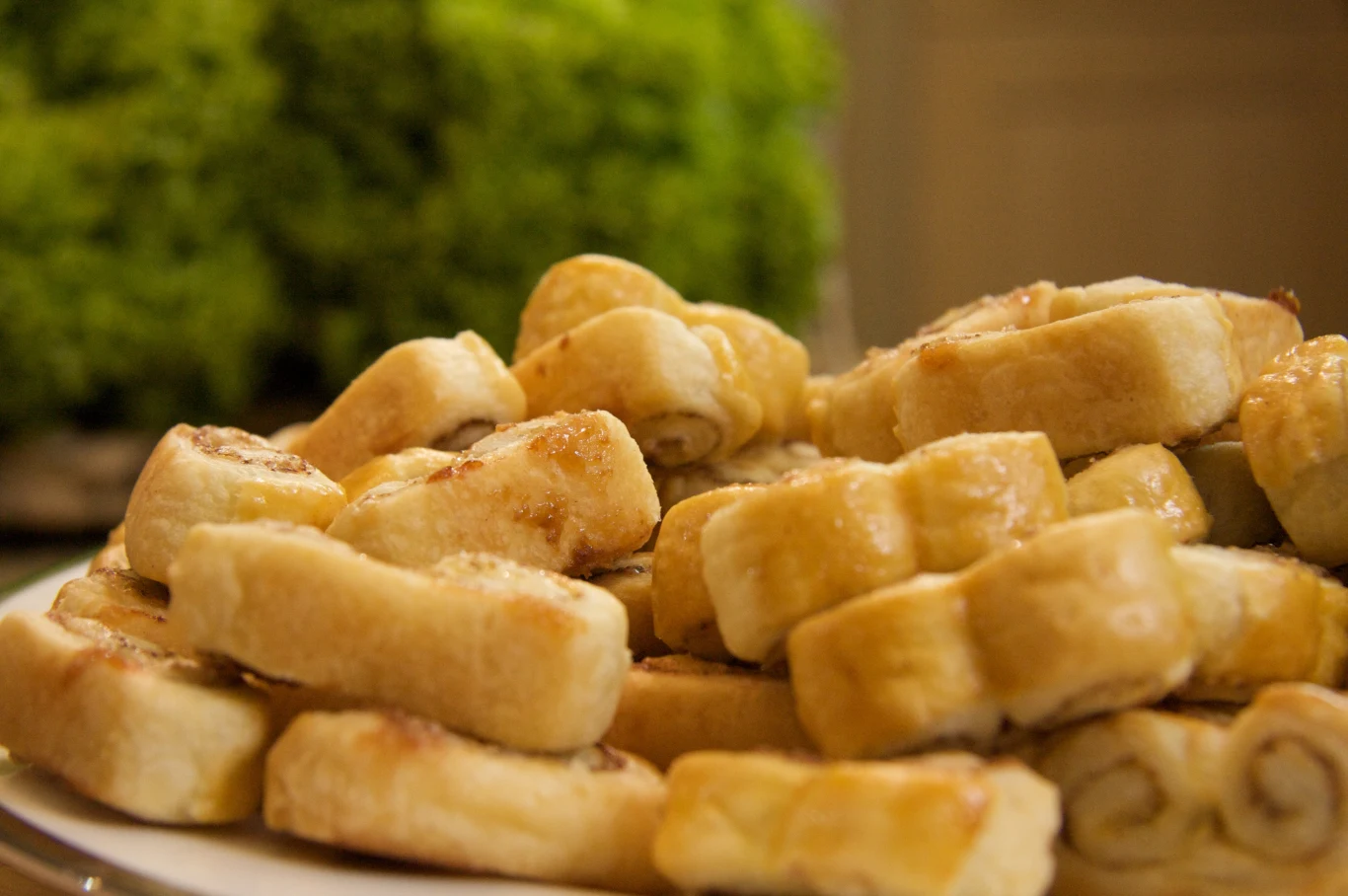 Stos maślanych rogalików ułożonych na białym talerzu, na pierwszym planie wypieki ze złocistą, połyskującą skórką, w tle zamazany zielony element, prawdopodobnie dekoracja lub roślina.