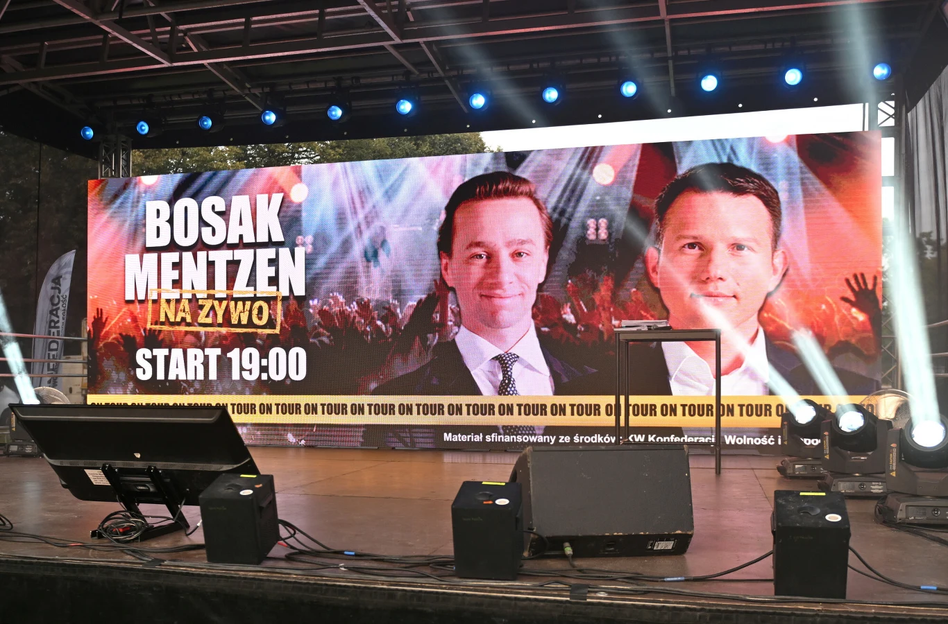 Liderzy Konfederacji praktycznie ze sobą nie rozmawiają Duży ekran sceniczny z wizerunkami dwóch mężczyzn i napisem informującym o wydarzeniu na żywo, na pierwszym planie elementy techniczne sceny takie jak statywy, monitory i światła.