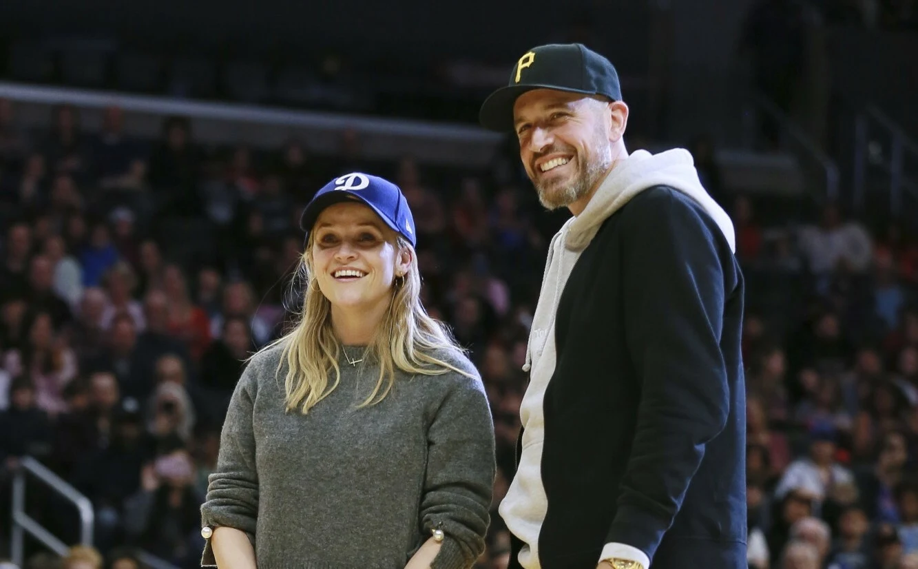 Reese Witherspoon i Jim Toth Dwie uśmiechnięte osoby stoją na tle tłumu w hali sportowej, obie noszą casualowe ubrania oraz czapki z daszkiem, mężczyzna ma zarost i jasną bluzę przewieszoną przez ramię.