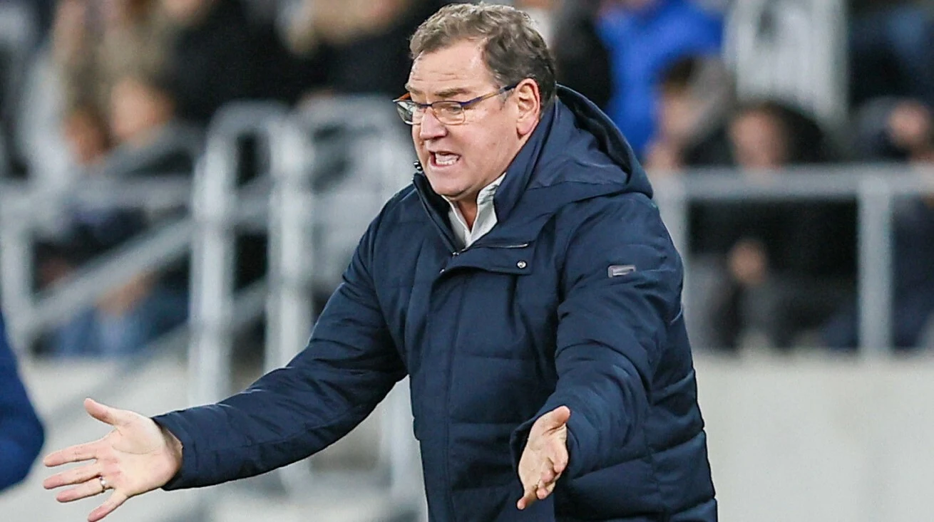 Mężczyzna w okularach ubrany w granatową kurtkę gestykuluje energicznie, stojąc na tle rozmytej trybuny stadionowej.