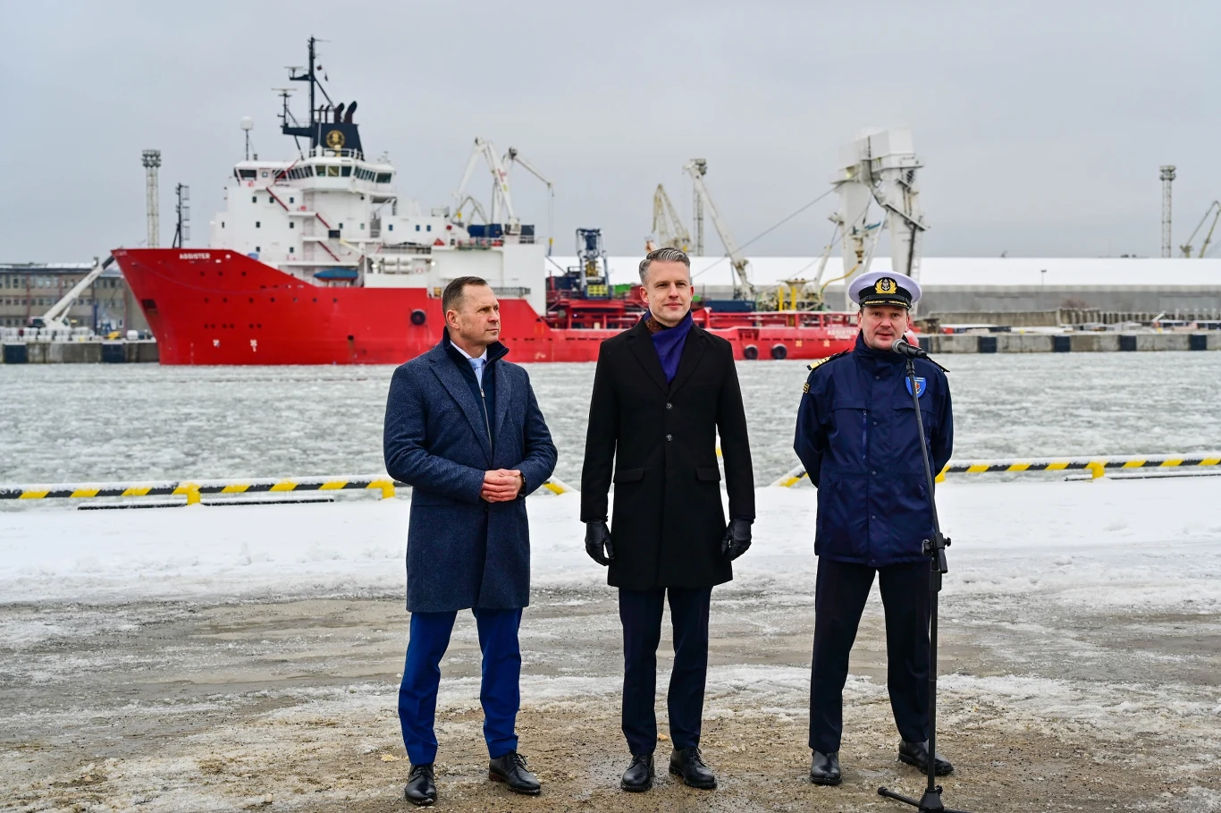 Ubiegły rok był dla polskich portów rekordowy pod względem przeładunku kontenerów Trzech mężczyzn stoi na tle portu morskiego, w pobliżu dużego czerwonego statku towarowego. Po prawej stronie znajduje się mężczyzna w mundurze i czapce kapitana, przed mikrofonem. W tle widoczne są dźwigi portowe, lód na wodzie oraz nowoczesna infrast...