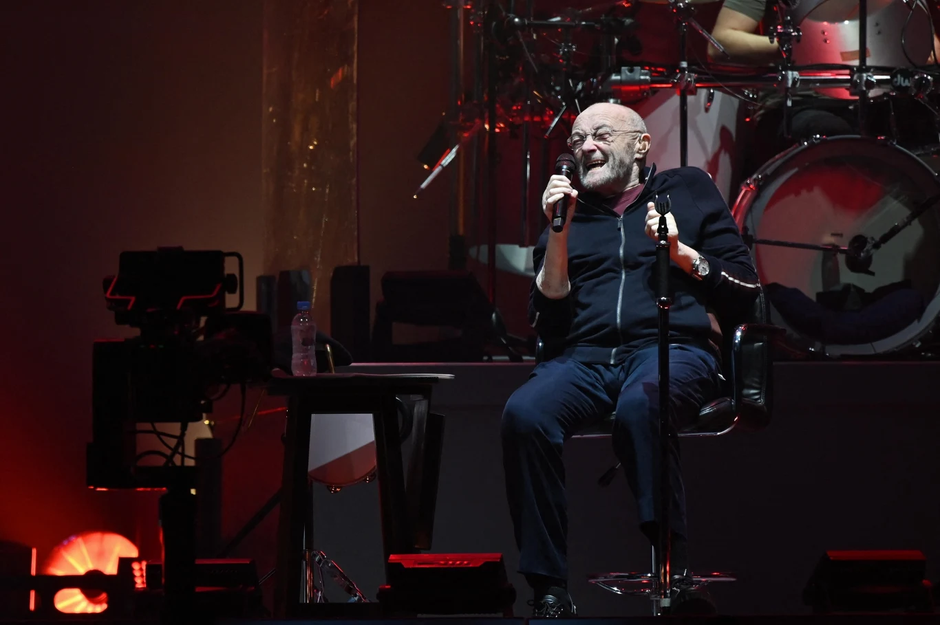 Starszy mężczyzna w ciemnym ubraniu siedzi na stołku na scenie z mikrofonem w obu rękach, śpiewając entuzjastycznie. W tle widoczna perkusja oraz reflektory sceniczne tworzące ciepłe, koncertowe oświetlenie.