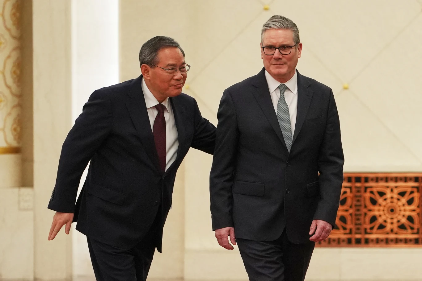 Chiński premier Li Qiang (L) i brytyjski premier Keir Starmer podczas spotkania w Pekinie Dwóch mężczyzn w eleganckich garniturach znajduje się w jasnym wnętrzu. Jeden z nich lekko dotyka pleców drugiego w geście uprzejmości. Tło stanowi zdobiona kratka wentylacyjna oraz klasyczna dekoracja ściany.