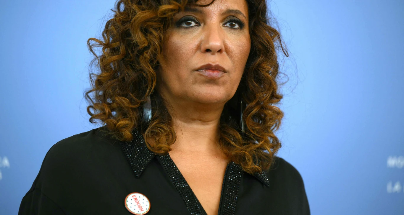 Kaouther Ben Hania na festiwalu filmowym w Wenecji 2025 Portret kobiety o kręconych włosach, ubranej w czarną koszulę z przypiętą okrągłą przypinką, na stonowanym niebieskim tle.