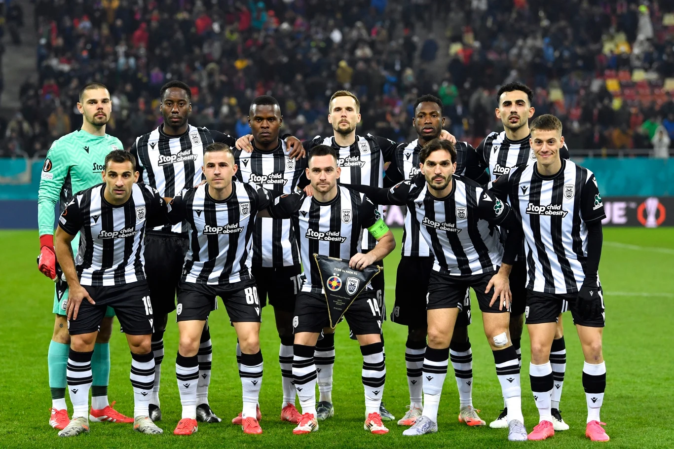 PAOK Saloniki Grupa jedenastu piłkarzy w czarno-białych strojach sportowych stoi razem na murawie stadionu, gotowa do rozpoczęcia meczu. W tle widać trybuny pełne kibiców.