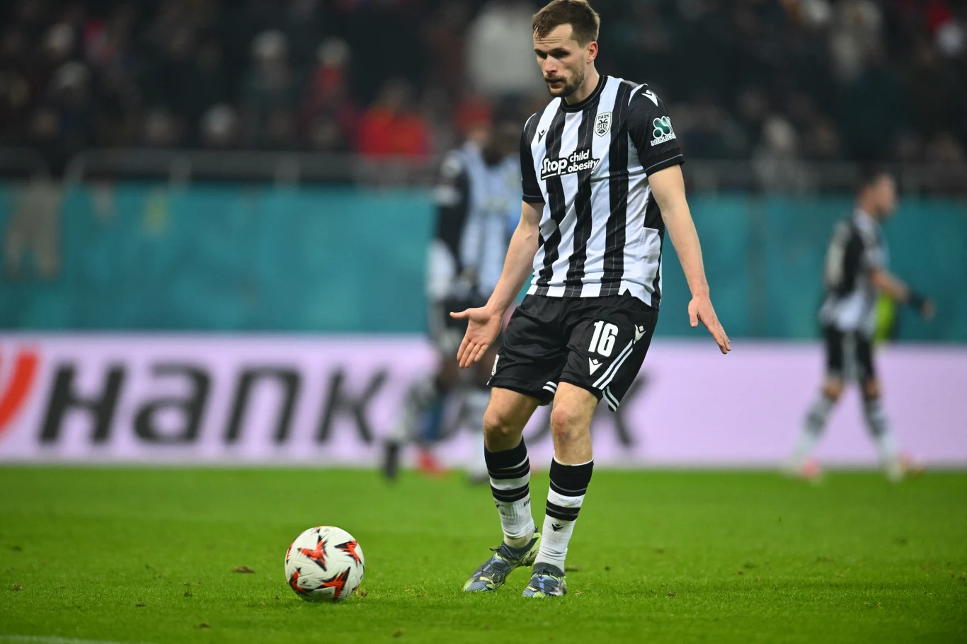 Tomasz Kędziora Piłkarz w czarno-białym stroju kontroluje piłkę na boisku podczas meczu. W tle widoczni inni zawodnicy i rozmyci kibice na trybunach.