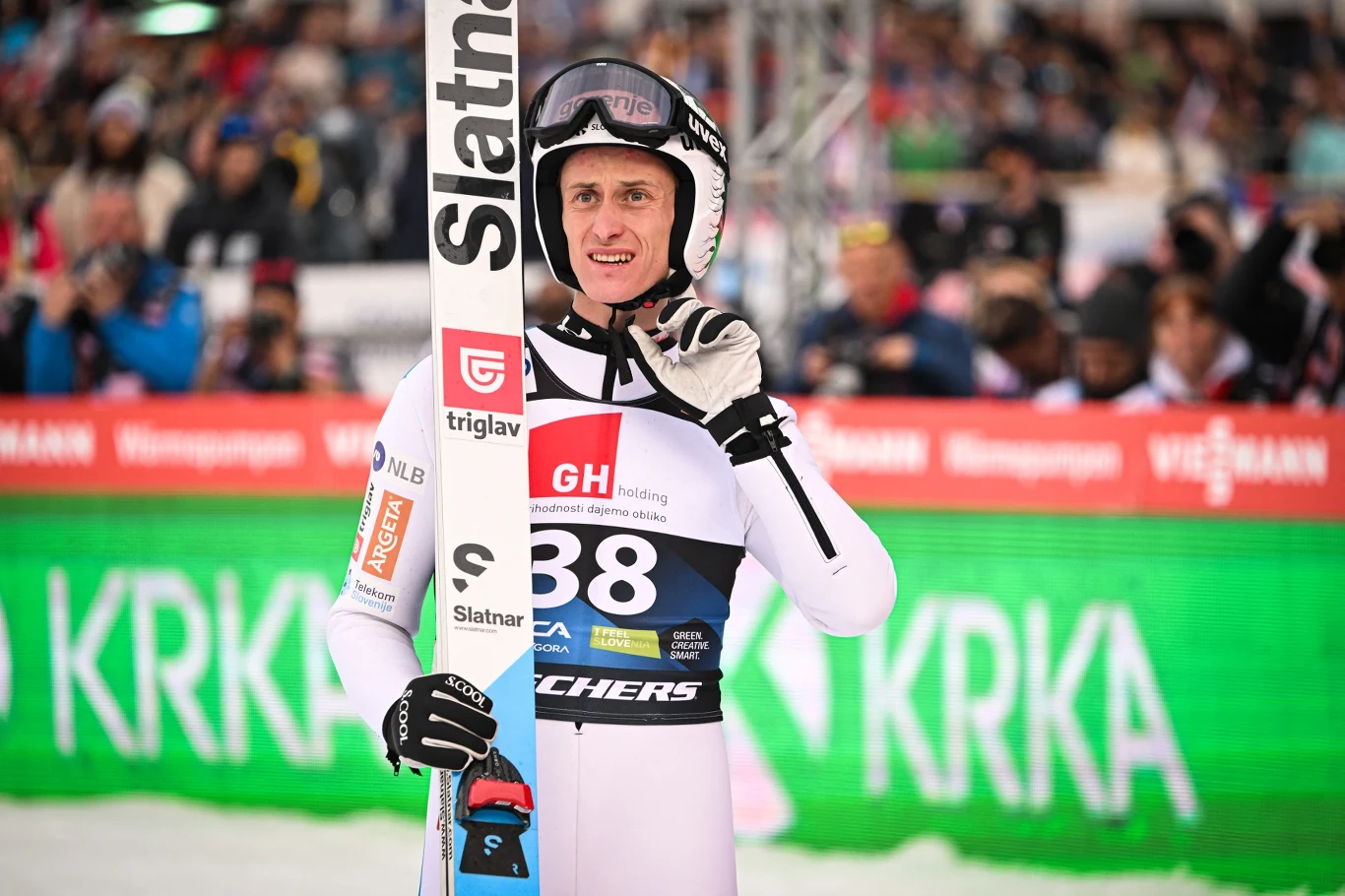 Skoczek narciarski w profesjonalnym kombinezonie i kasku trzyma narty, w tle kibice na trybunach oraz widoczne są reklamy sponsorów i zielony baner KRKA.