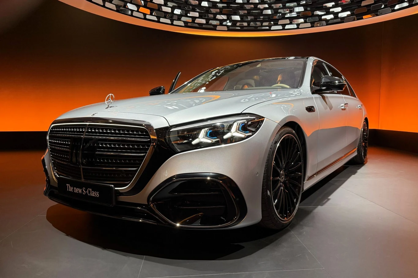 Mercedes klasy S przeszedł modernizację. Zakres zmian robi wrażenie. Luksusowy samochód Mercedes-Benz klasy S w kolorze srebrnym ustawiony na wystawie, z podświetloną pomarańczową ścianą w tle, wyeksponowany z przodu, wyróżniający się nowoczesnym grillem, dużymi felgami i eleganckim wykończeniem.