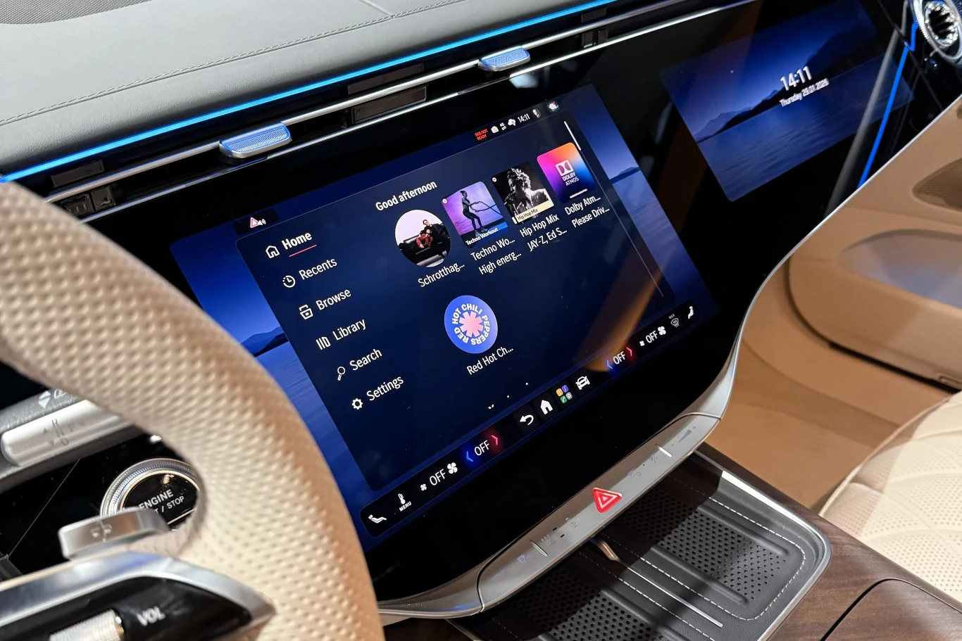 Główne miejsce zajmuje 14,4-calowy ekran multimediów. Znajdujące się pod spodem przełączniki są fizyczne, ale np. głośnością multimediów możemy sterować również przesuwając po przełączniku. Nowoczesny kokpit samochodu wyposażony w duży ekran dotykowy z interfejsem multimedialnym, otoczony elegancką tapicerką oraz elementami wykończenia w jasnych kolorach, fragment kierownicy widoczny z lewej strony.