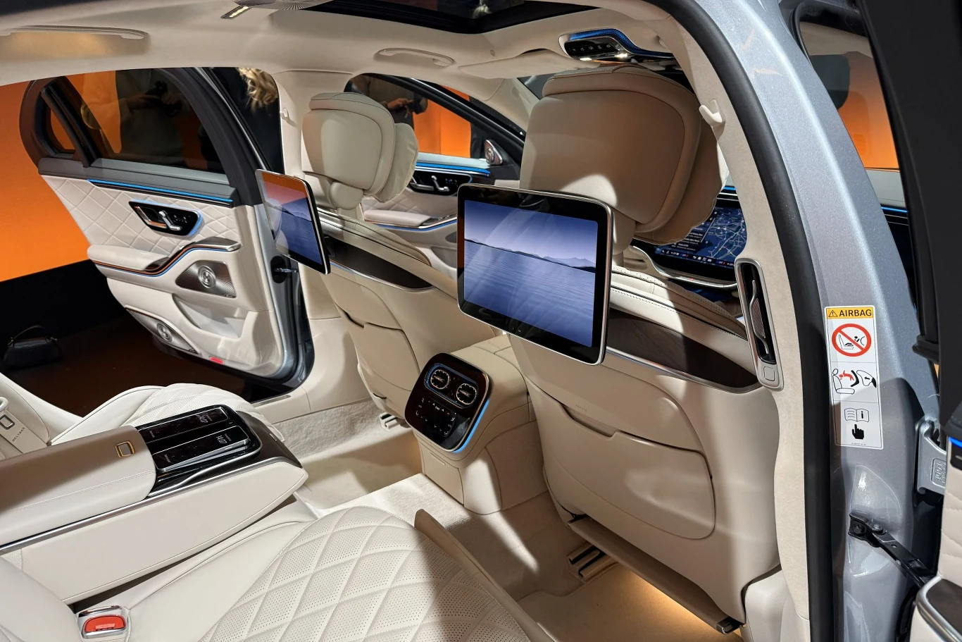 Z tyłu znajdziemy teraz dwa większe 13,1-calowe ekrany. Luksusowe wnętrze samochodu z jasną, skórzaną tapicerką, dużą ilością miejsca na nogi i dwoma ekranami multimedialnymi zamontowanymi z tyłu przednich foteli. Centralna konsola rozdzielająca tylne siedzenia wyposażona w panel sterowania. Wyraźnie widocz...