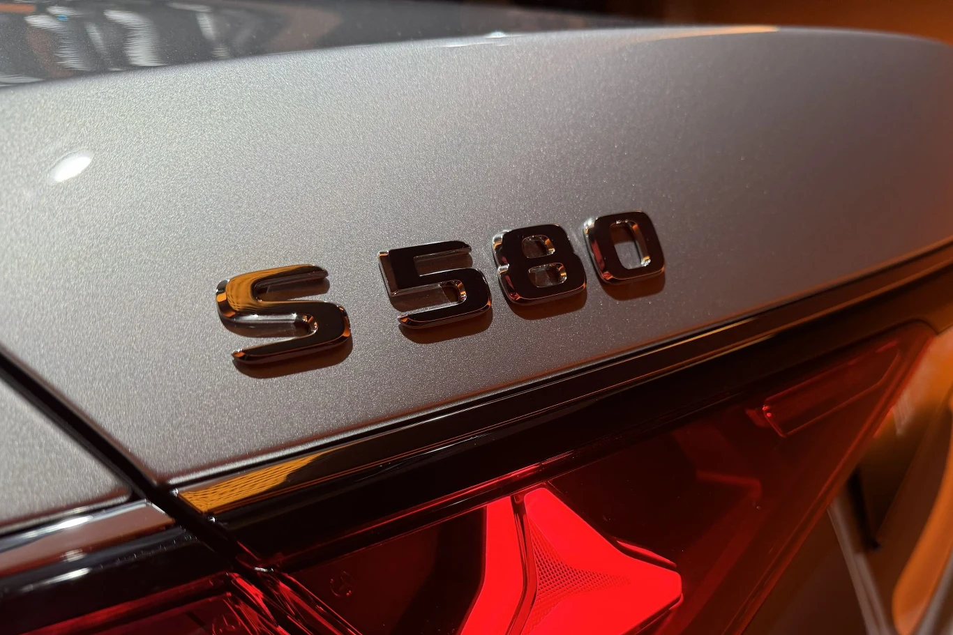 Zarówno w przypadku wariantów benzynowych, jak i wysokoprężnych silnik będzie wspomagać 23-konny rozruszniko-alternator. Chromowany emblemat S 580 umieszczony na srebrnej karoserii luksusowego samochodu z widocznym fragmentem tylnej lampy o czerwonym podświetleniu.