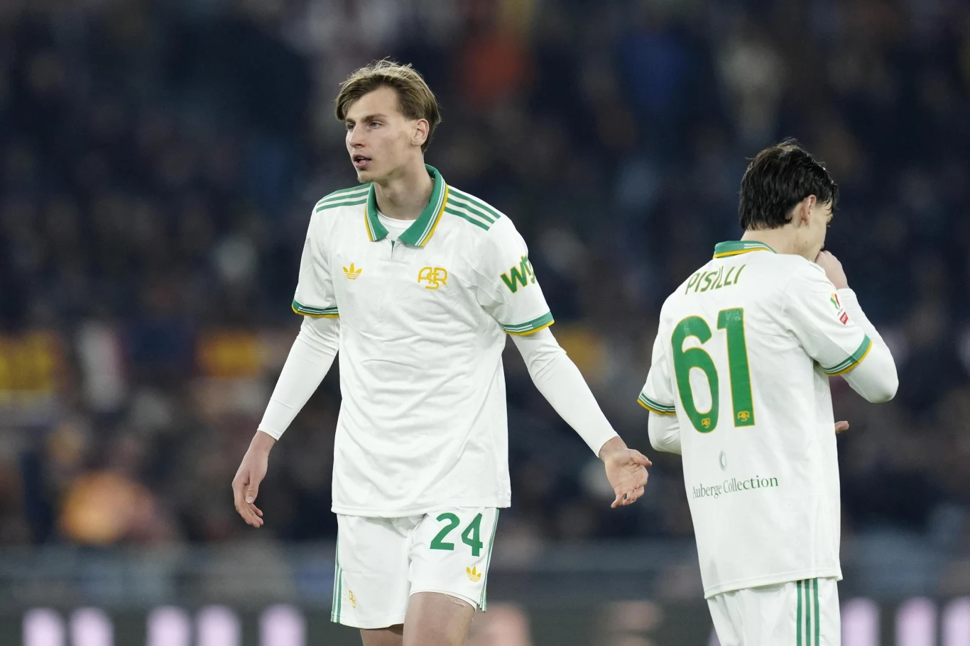 Dwóch piłkarzy w białych strojach z zielonymi akcentami stoi na murawie stadionu, jeden z nich ma widoczny numer 24, drugi 61, a w tle rozmyta publiczność.