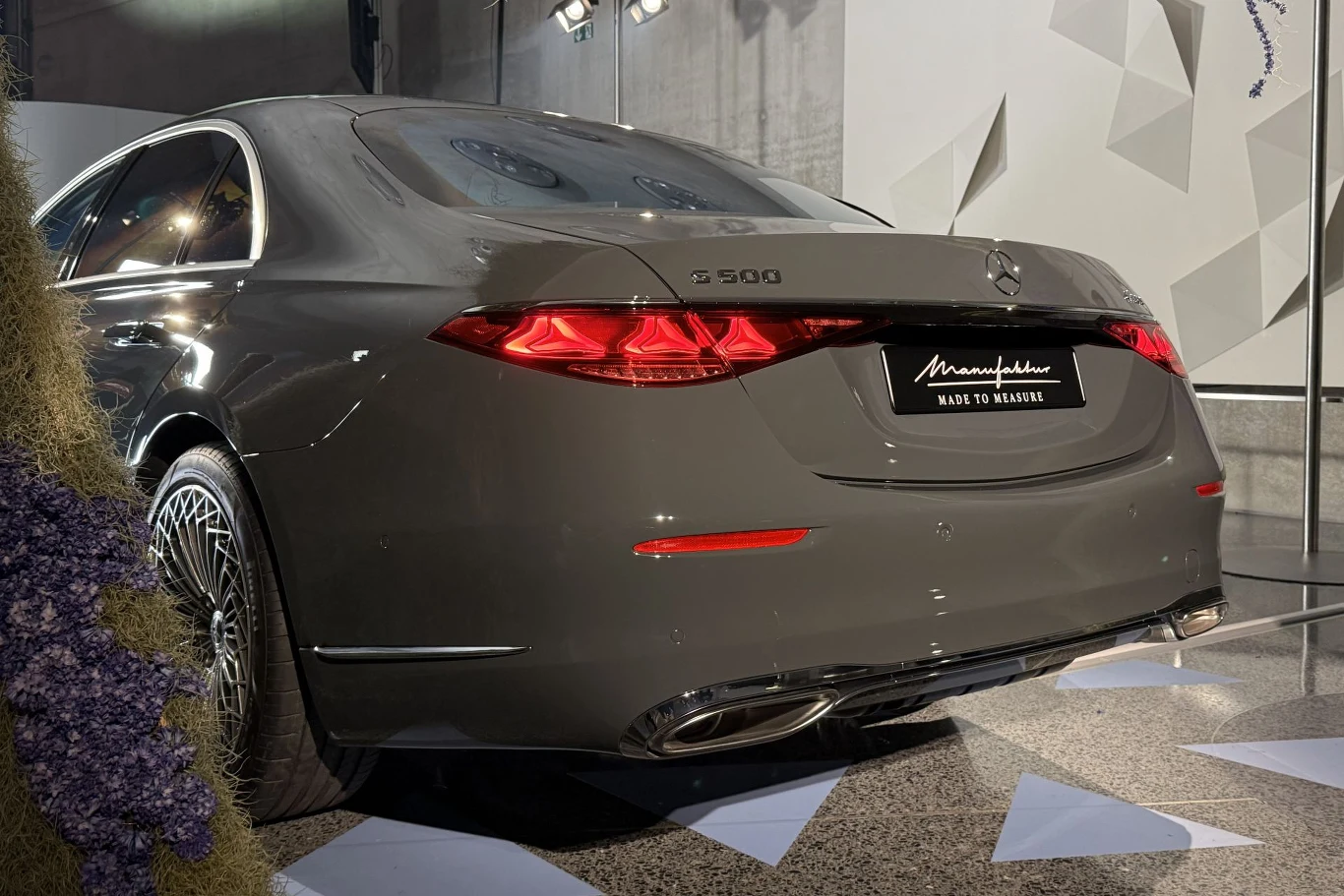 Mercedes klasy S po liftingu wciąż będzie dostępny w ramach programu personalizacji MANUFAKTUR. Szary Mercedes-Benz S500 w wersji Maybach prezentowany na wystawie, widok od tyłu, widoczne eleganckie detale karoserii, charakterystyczne tylne lampy, elementy wystroju wnętrza ekspozycji oraz geometryczne wzory na ścianie.