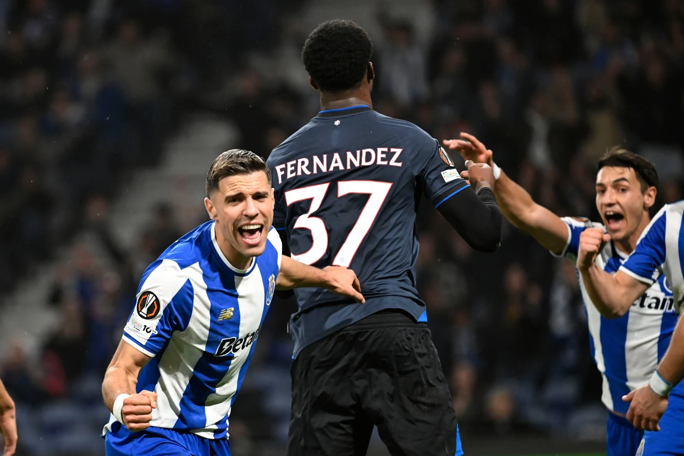 Radość piłkarza drużyny ubranej w niebiesko-białe pasy po zdobyciu bramki podczas meczu piłki nożnej, zawodnik w czarno-niebieskiej koszulce z nazwiskiem Fernandez z tyłu, kibice i inni zawodnicy w tle.