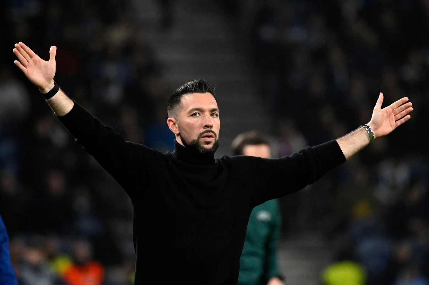 Mężczyzna z zarostem w czarnym golfie stoi z uniesionymi rękami na tle rozmytej publiczności na stadionie, wygląda na zaskoczonego lub niezadowolonego, wokół niego znajdują się inne osoby, ale są one nieostre i w tle.