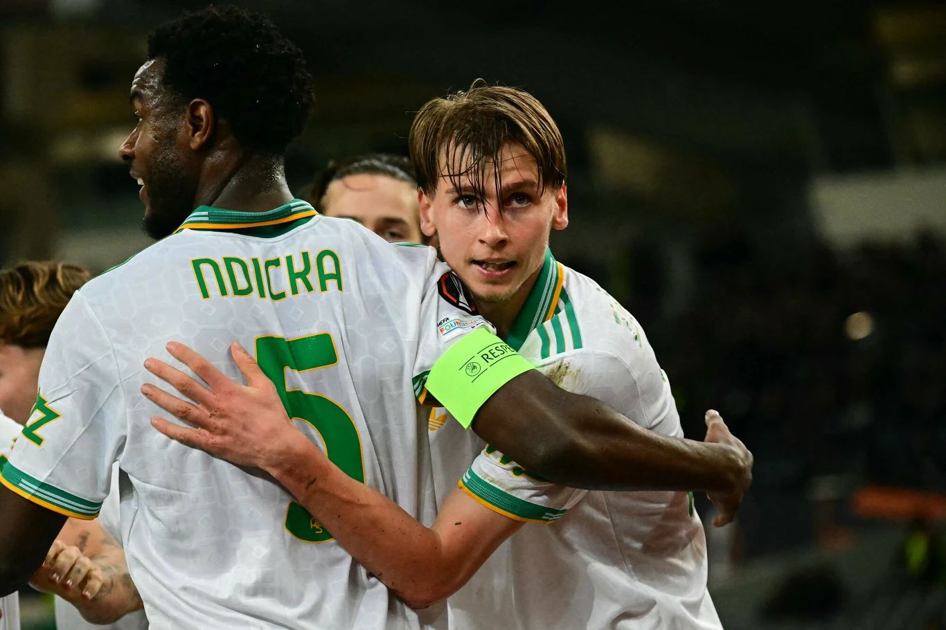 Zawodnicy drużyny piłkarskiej w białych koszulkach celebrują wspólnie zdobycie bramki, jeden z nich obejmuje kolegę z zespołu, w tle widoczny stadion.