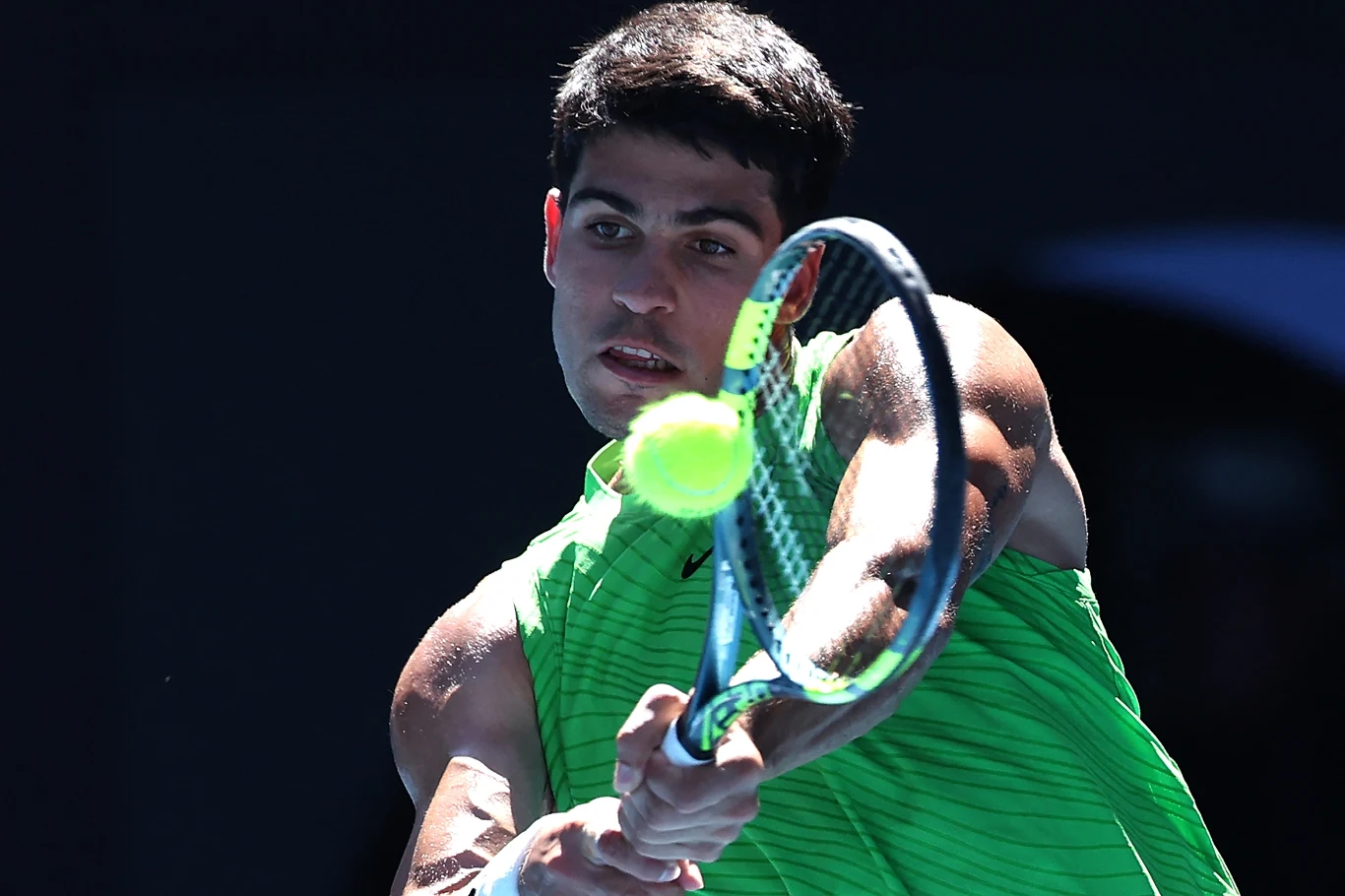 Młody mężczyzna w intensywnie zielonej koszulce uderza piłkę tenisową rakietą, skupiony na grze, rozświetlony światłem, tło ciemne.