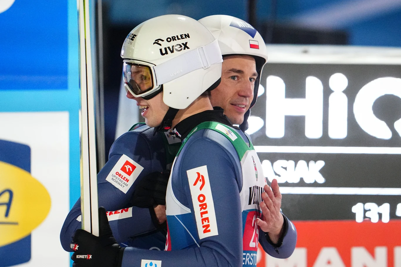 Kacper Tomasiak i Kamil Stoch powalczą o medale na zimowych igrzyskach olimpijskich 2026 Dwóch skoczków narciarskich w kombinezonach i kaskach, obejmujących się na tle tablicy wyników z widocznymi logotypami sponsorów.