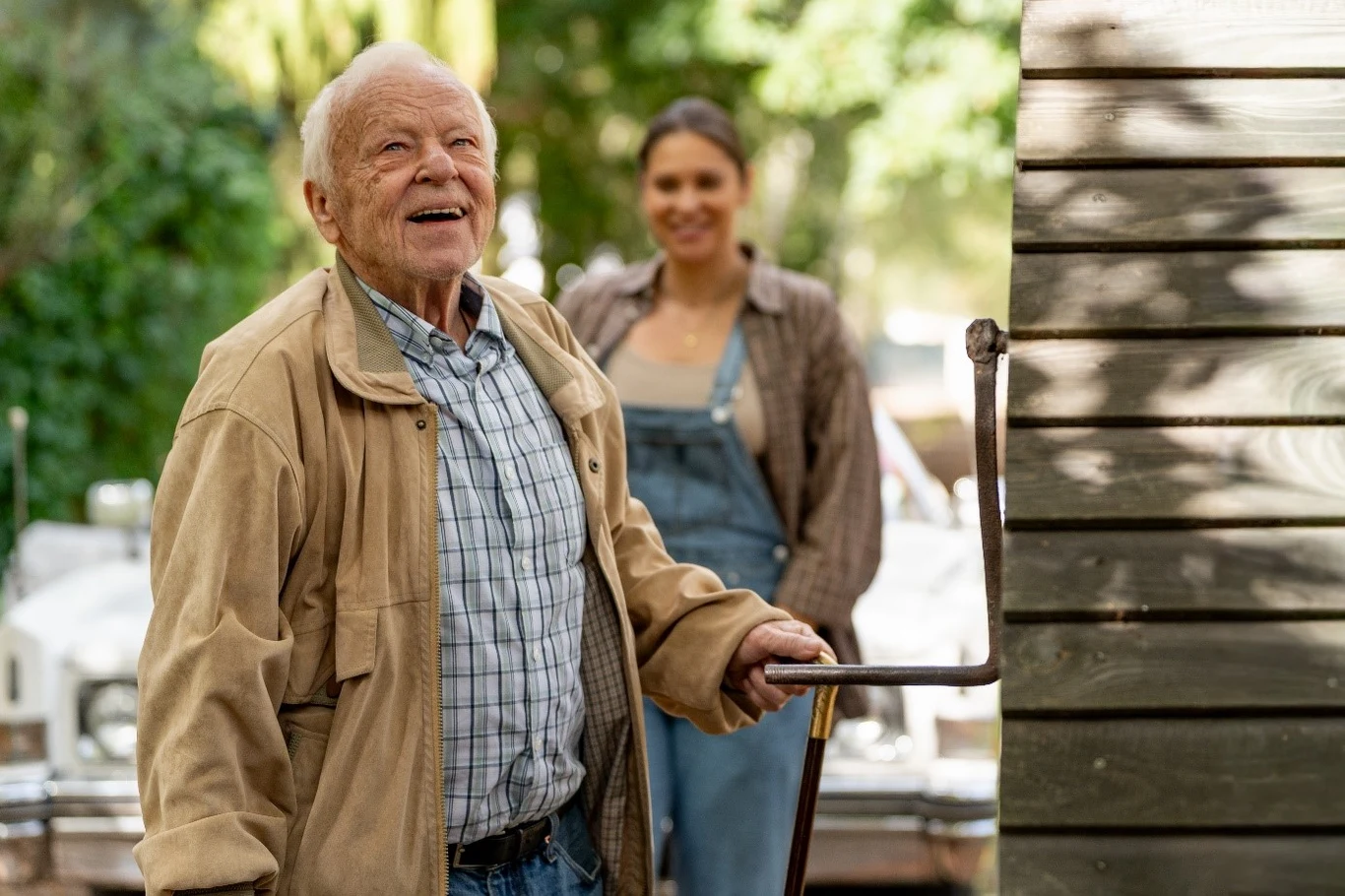 Marian Opania Starszy mężczyzna w jasnej kurtce i koszuli trzyma metalową laskę, stoi na zewnątrz obok drewnianej ściany budynku. W tle młoda kobieta w ogrodniczkach uśmiecha się, na drugim planie rozmyta, zielona roślinność.