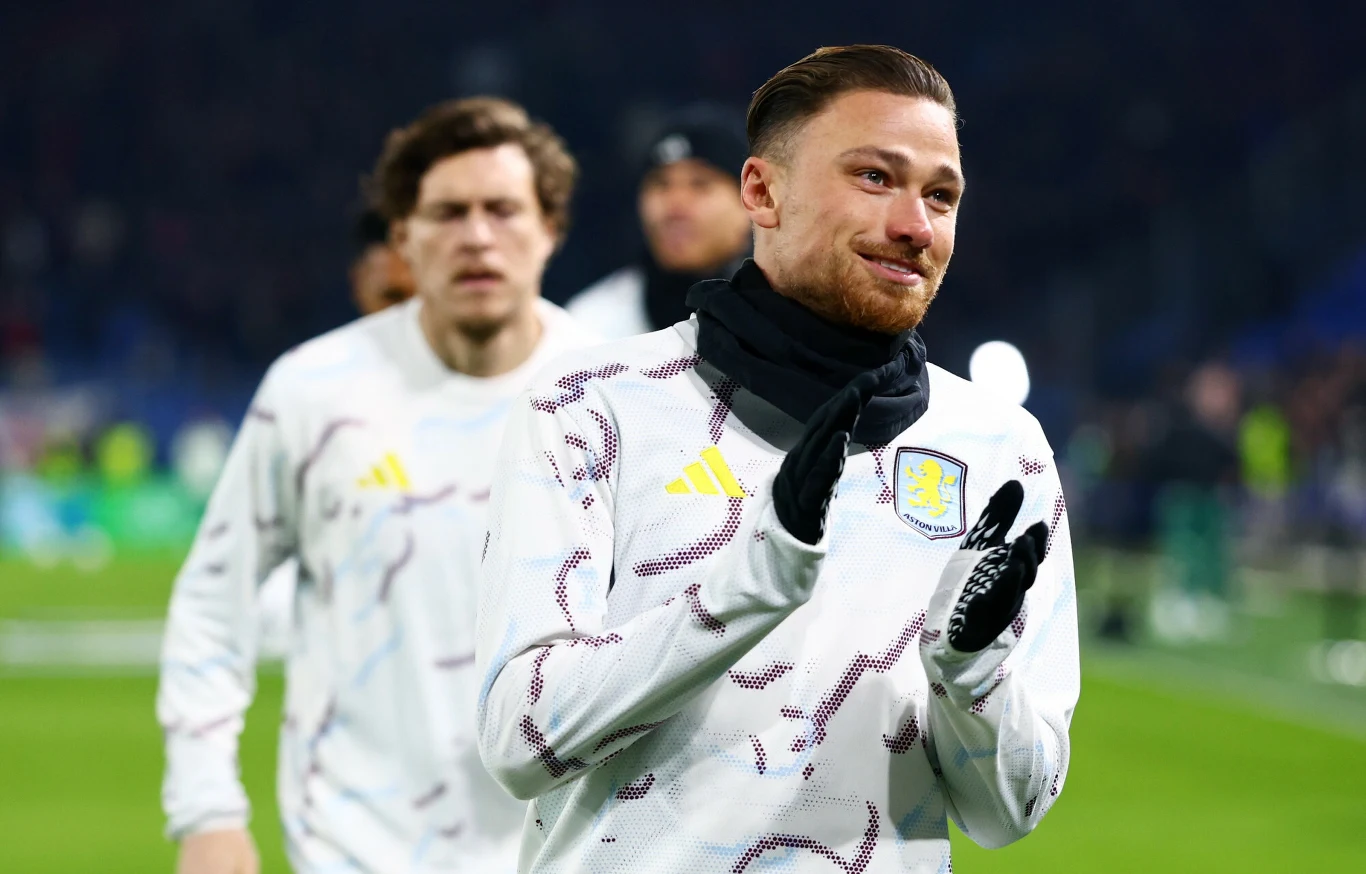 Piłkarz w białej bluzie treningowej z herbem klubu Aston Villa na piersi, zaciśnięte rękawiczki na dłoniach, uśmiechnięty, w tle drugi zawodnik rozgrzewający się na boisku.