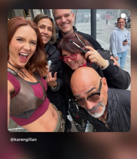 Grupa pięciu uśmiechniętych osób pozuje do selfie na ulicy, wśród nich kobieta w kostiumie na pierwszym planie oraz kilka osób z akcesoriami fryzjerskimi i makijażu, w tle przechodzień i miejskie otoczenie.