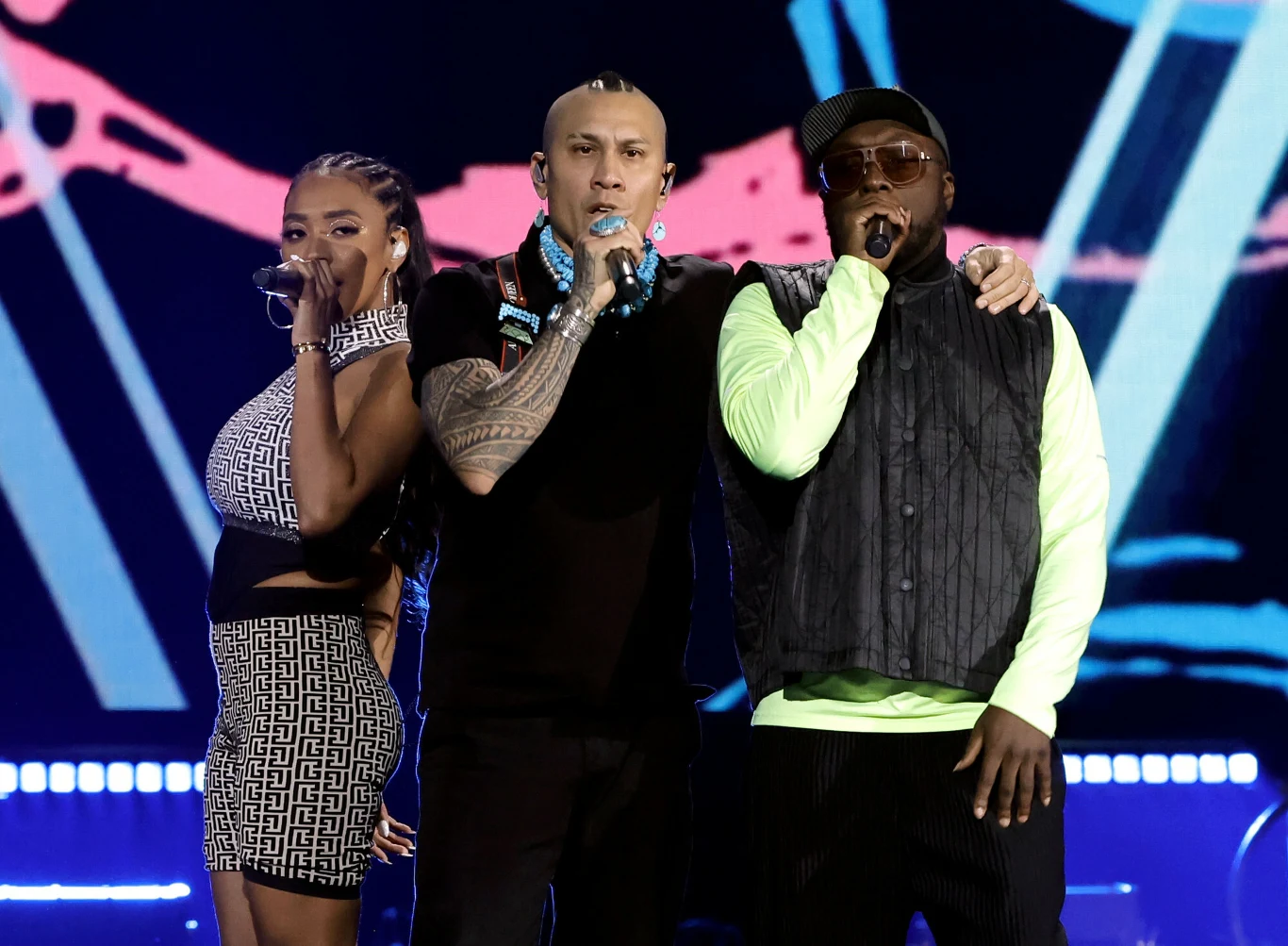 Black Eyed Peas w czerwcu wystąpią w Polsce Trzech wykonawców stojących na scenie, śpiewających do mikrofonów, z tłem scenicznym w neonowych kolorach i światłami podkreślającymi występ muzyczny.
