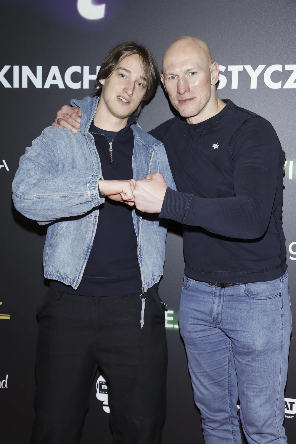 Dwóch mężczyzn stoi obok siebie na tle planszy promocyjnej, wykonując wspólny gest pięści, co sugeruje wzajemny szacunek lub przyjaźń. Jeden z nich ubrany jest w jasnoniebieską kurtkę dżinsową, drugi w ciemnoniebieski sweter i dżinsy.