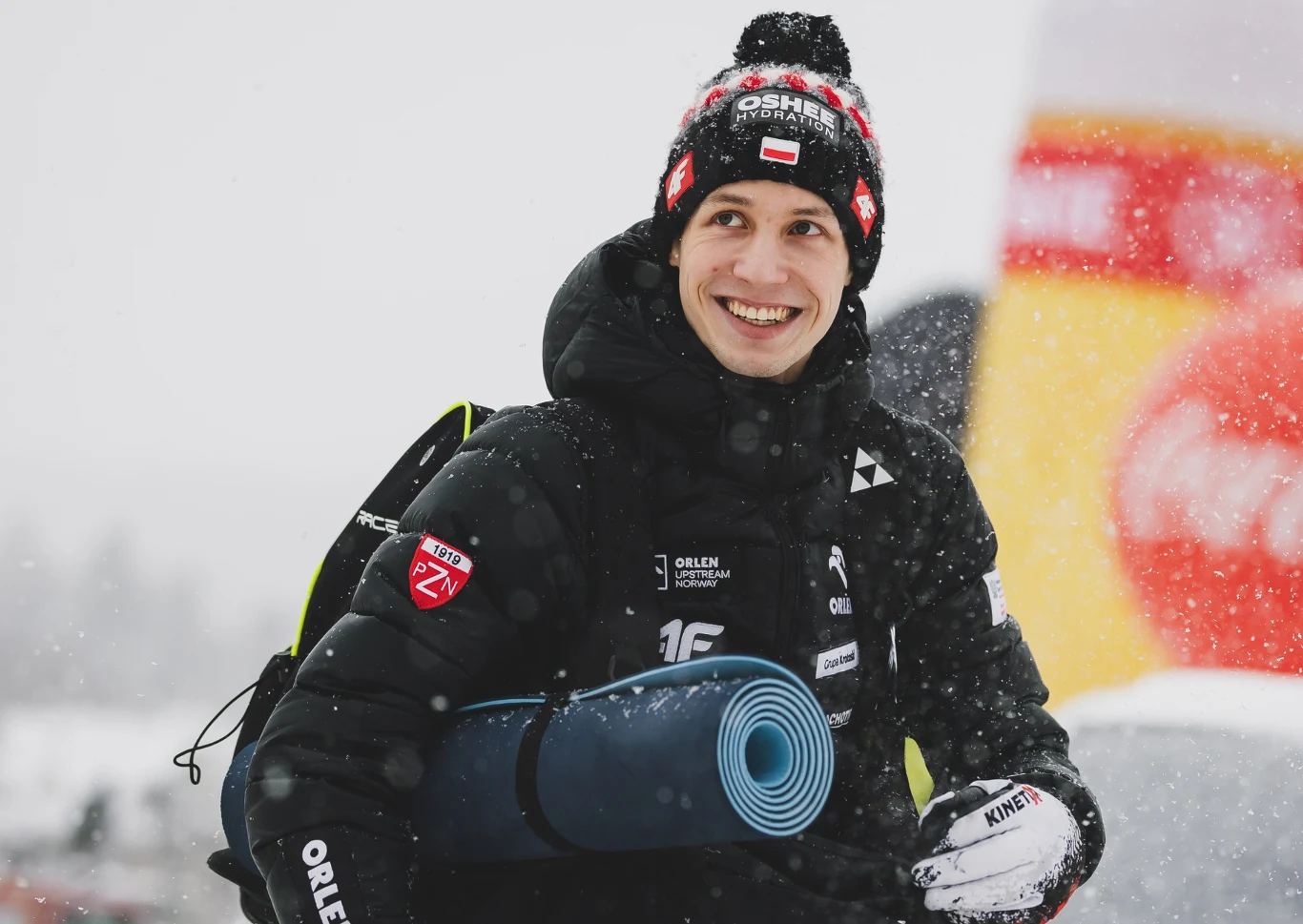 Uśmiechnięty mężczyzna w zimowej kurtce i czapce z napisem, trzymający zrolowaną matę sportową, w tle śnieg oraz kolorowy balon reklamowy.