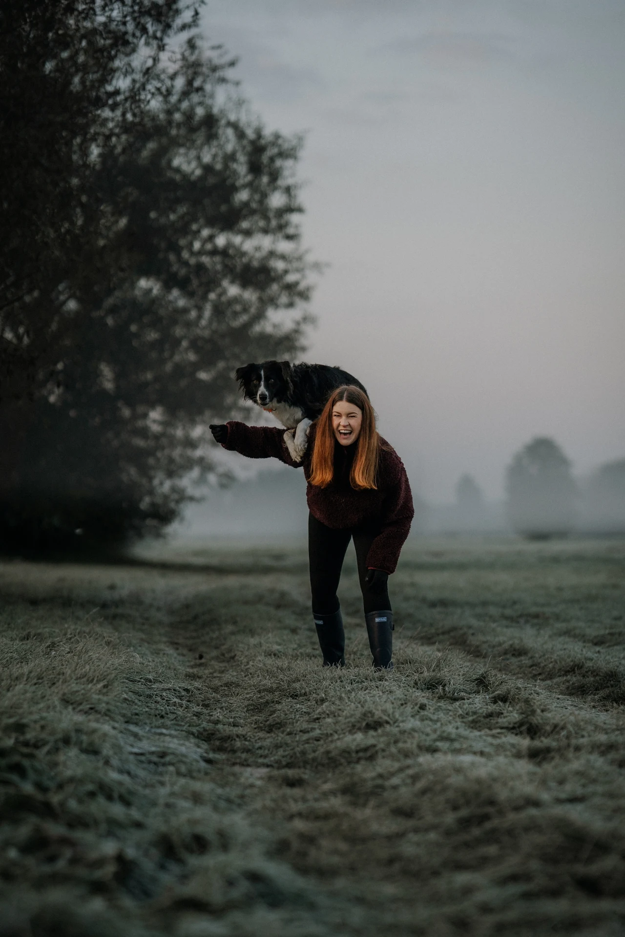 Młoda kobieta w kaloszach idzie przez zamglone pole o poranku, trzymając czarno-białego psa na ramionach. W tle widoczne drzewa i delikatna mgła.