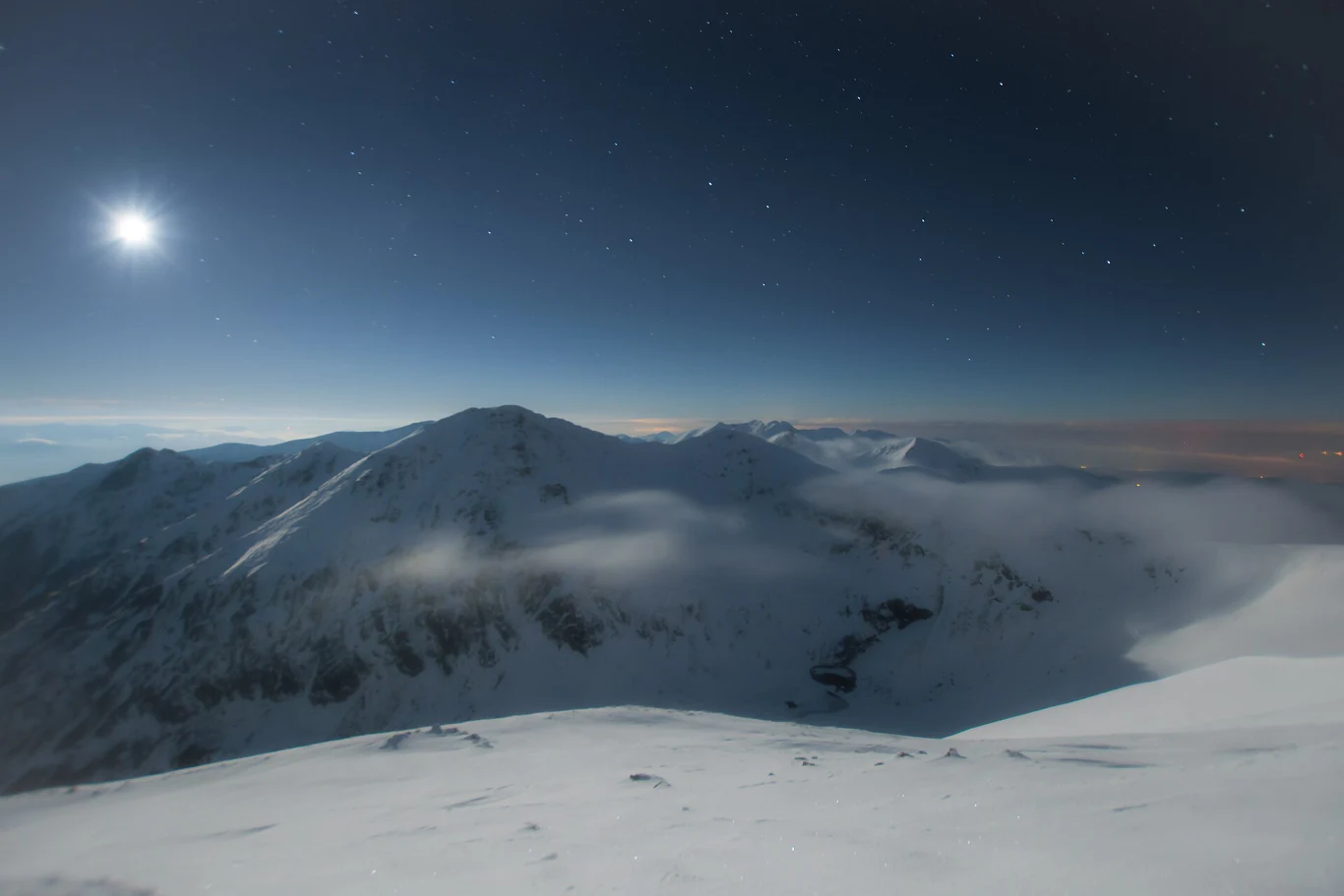 Ośnieżone szczyty gór rozciągają się pod ciemnym, gwiaździstym niebem z jasno świecącym księżycem, a nad górami unoszą się delikatne mgły tworząc spokojną, nocną atmosferę.