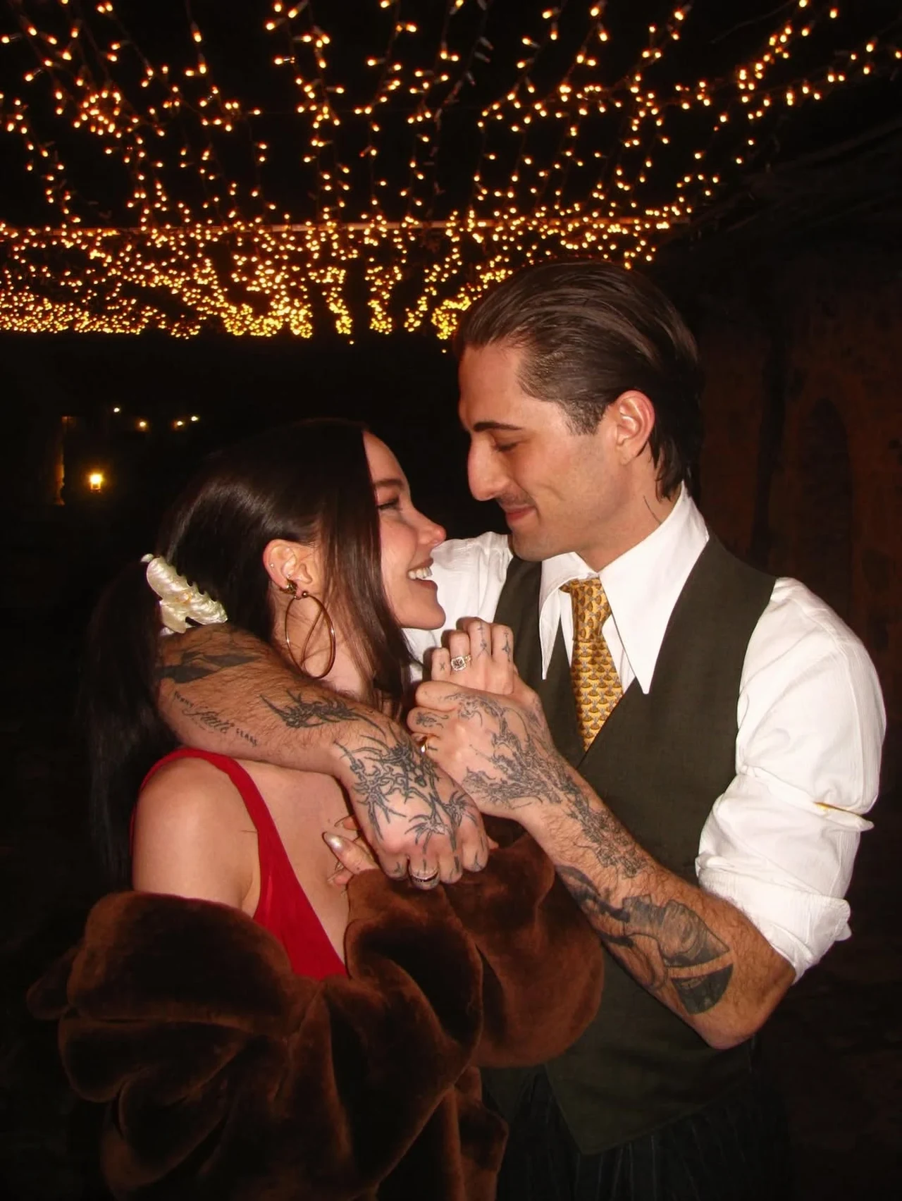 Dove Cameron i Damiano David zaręczyli się Para obejmująca się w romantycznej atmosferze pod girlandami świateł, mężczyzna ubrany elegancko z tatuażami na rękach, kobieta w czerwonej sukience i brązowym futrze, oboje uśmiechnięci, patrzą sobie w oczy.