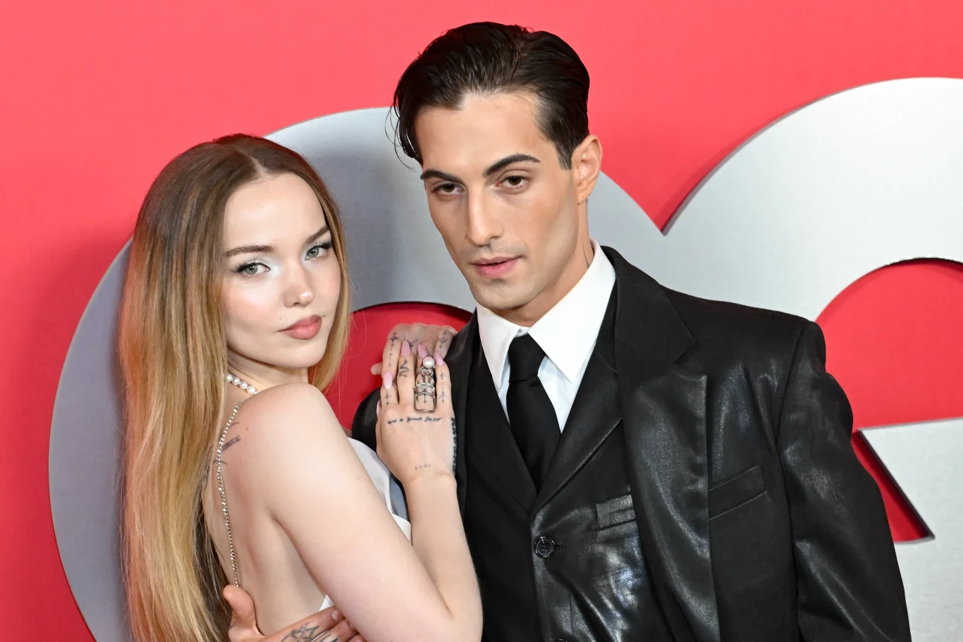 Dove Cameron i Damiano David Dwie osoby pozują razem przy czerwonym tle z dużą literą GQ, kobieta w jasnej sukni z długimi, prostymi włosami i mężczyzna w czarnym garniturze z białą koszulą i czarnym krawatem. Kobieta opiera rękę na ramieniu mężczyzny, oboje patrzą prosto w obiektyw.