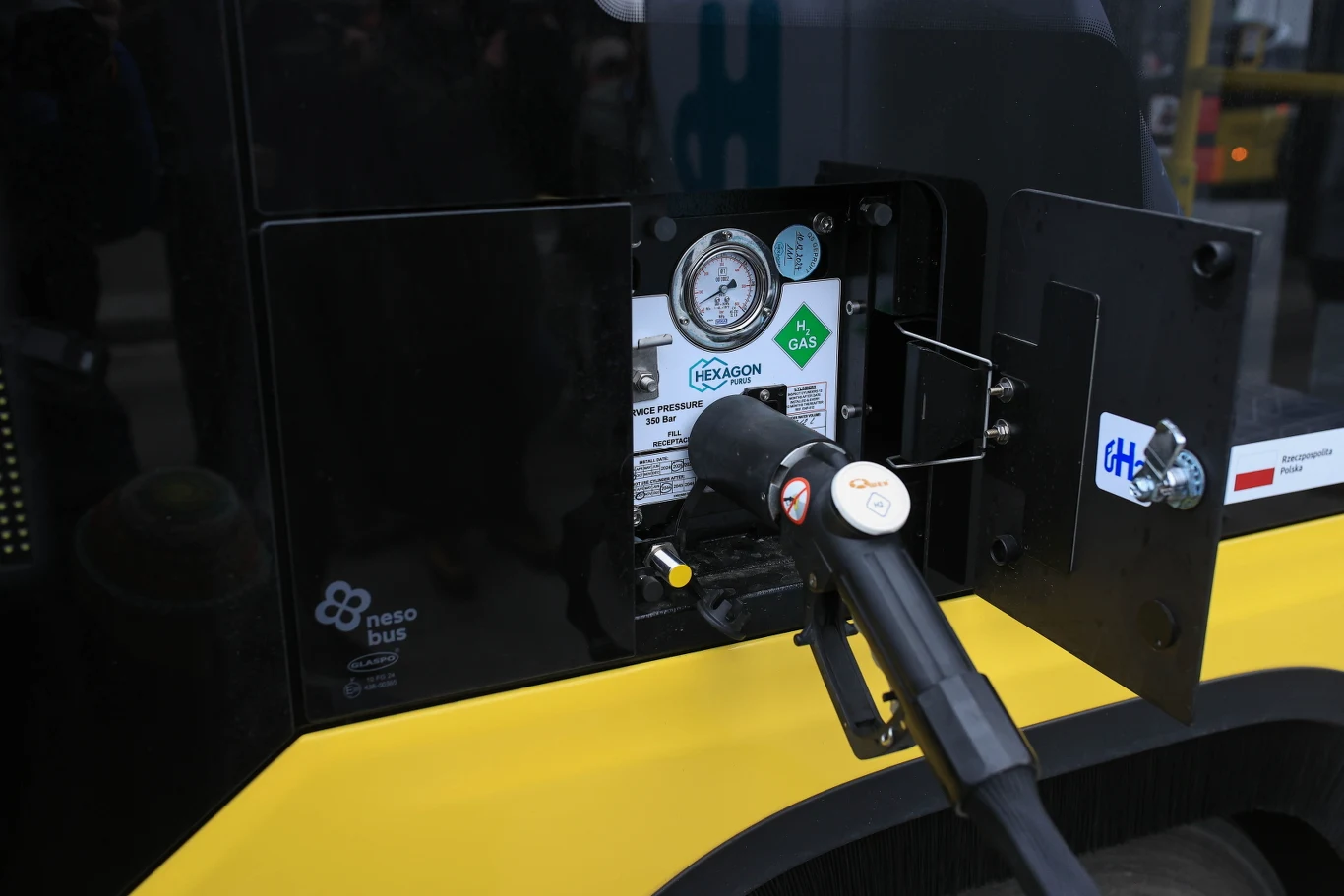 Stacja tankowania wodoru z widocznym pistoletem podpiętym do zbiornika autobusu, wskaźnikiem ciśnienia oraz oznaczeniami informującymi o rodzaju gazu.