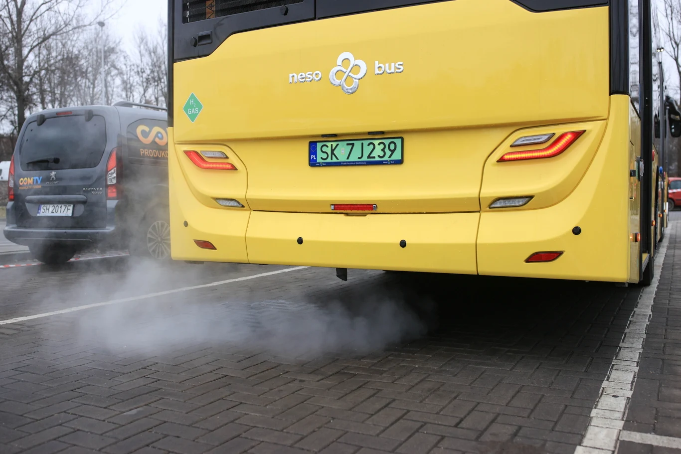 Żółty autobus emituje spaliny z rury wydechowej na parkingu, w tle widoczny zaparkowany samochód osobowy oraz drzewa bez liści.