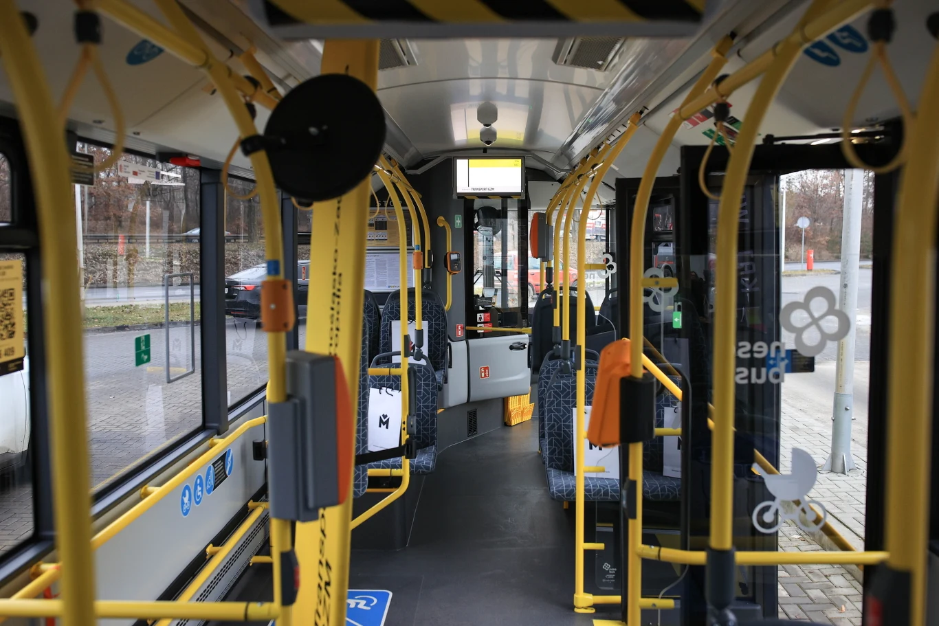 Wnętrze miejskiego autobusu z widocznymi miejscami siedzącymi, uchwytami dla pasażerów, żółtymi poręczami i dużymi oknami po bokach, zza których widać fragment ulicy i przystanku.