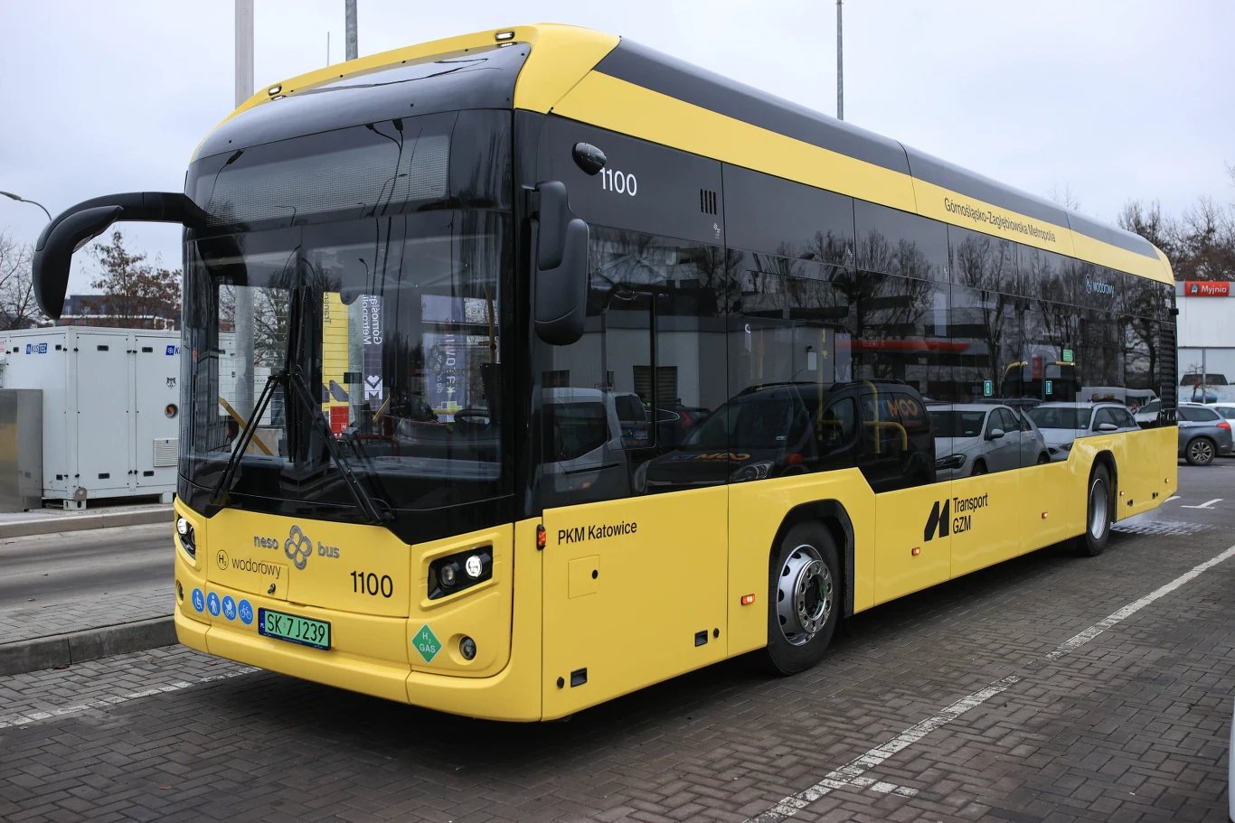 Nowoczesny, żółty autobus elektryczny stojący na przystanku w miejskim otoczeniu, obok innych samochodów, z wyraźnie widocznym oznaczeniem linii oraz logo przewoźnika komunikacji miejskiej.