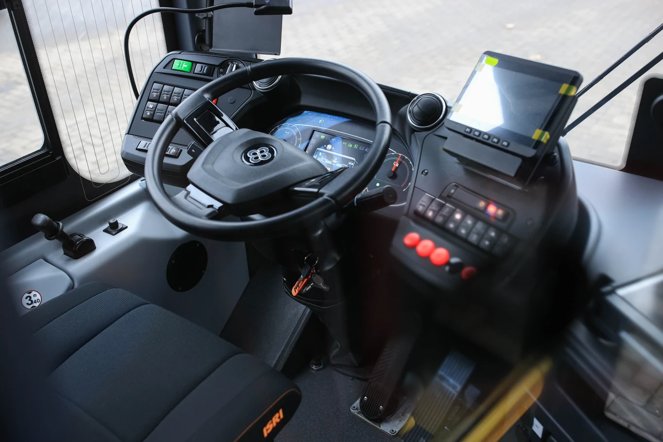 Kabina kierowcy autobusu z widoczną kierownicą, cyfrowym panelem sterowania, monitorem oraz zestawem przycisków i przełączników. Miejsce siedzące kierowcy obite ciemnym materiałem.