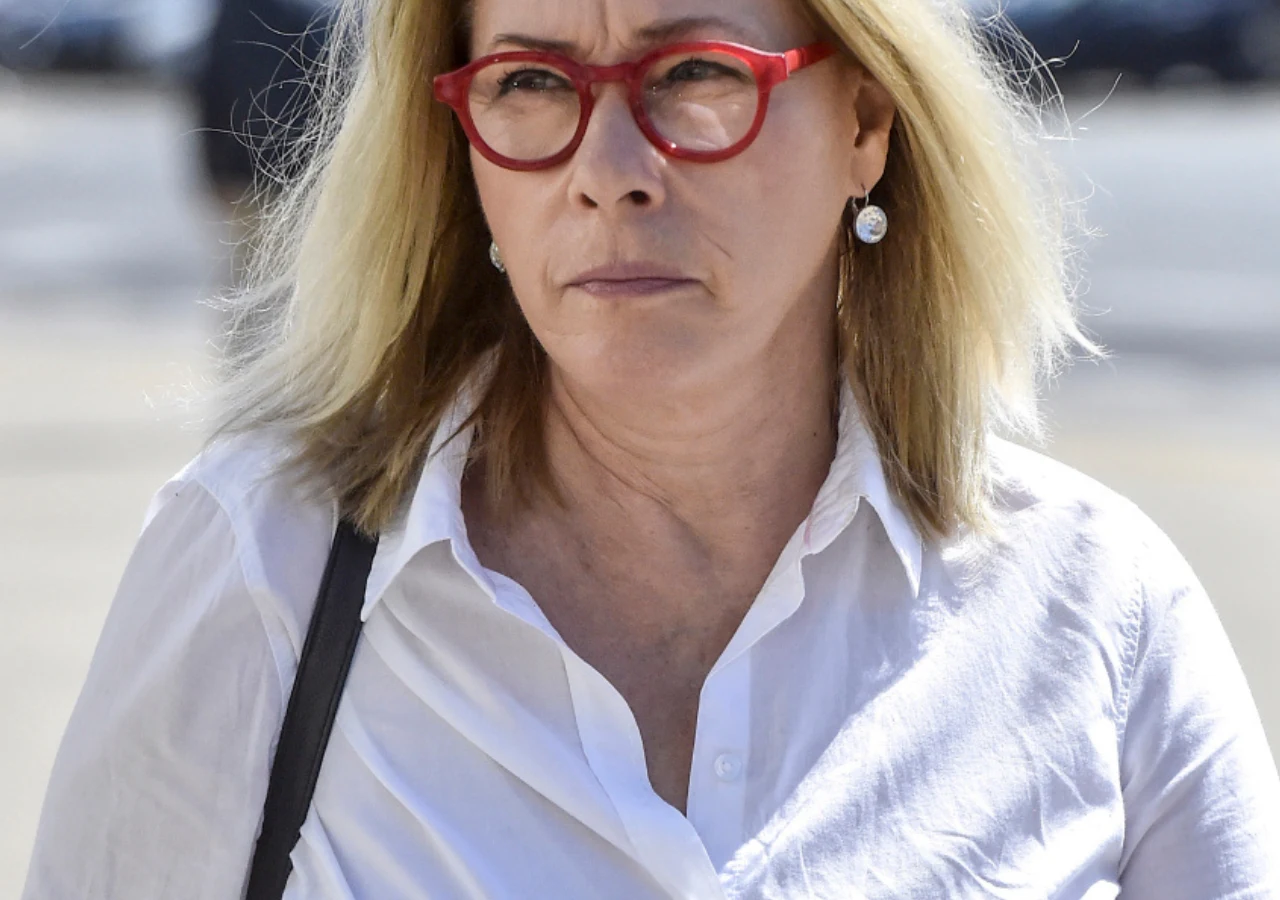 Małgorzata Potocka Kobieta w okularach o czerwonych oprawkach, ubrana w białą koszulę, z jasnymi włosami, idzie ulicą w świetle dziennym. Jej wyraz twarzy jest poważny, a w tle widoczne są rozmyte sylwetki samochodów.