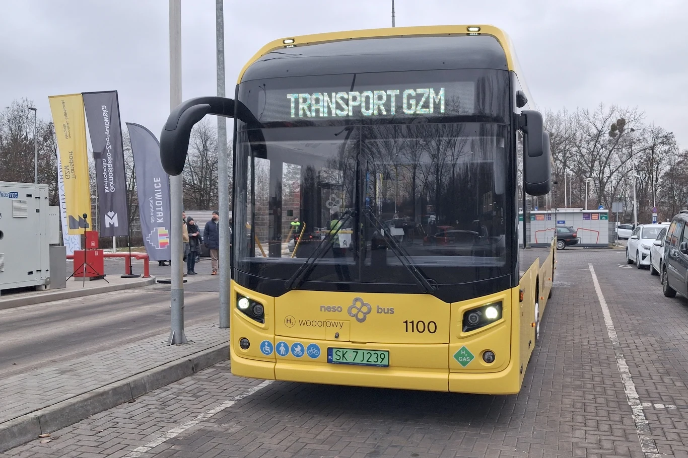 Żółty autobus miejski z napisem TRANSPORT GZM na tablicy kierunkowej stoi na przystanku, w tle widoczne są samochody, znaki drogowe oraz drzewa bez liści.