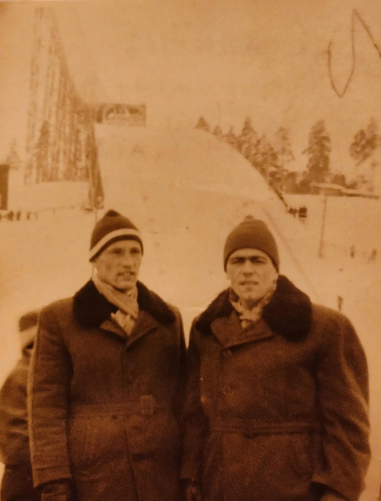 Dwóch mężczyzn w zimowych płaszczach i czapkach stoi na tle dużej skoczni narciarskiej, w otoczeniu zaśnieżonego krajobrazu i drzew widocznych w oddali.