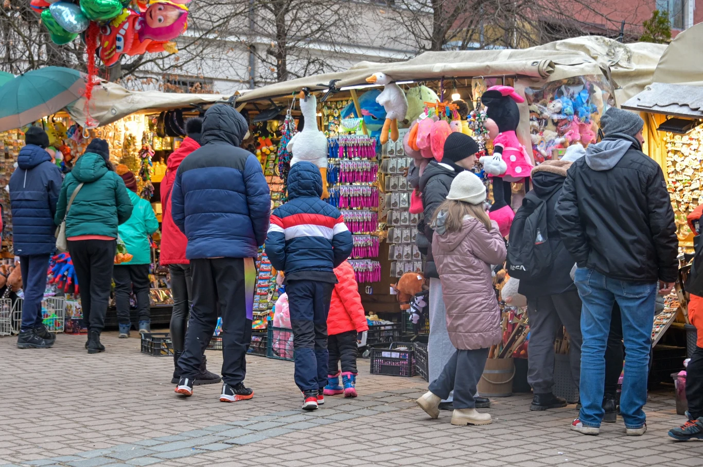 Ludzie ubrani w zimowe kurtki stoją przy stoiskach jarmarcznych pełnych kolorowych zabawek, maskotek oraz balonów. Na pierwszym planie dzieci oglądają różnorodne produkty, a w tle widać dekoracje i inne osoby odwiedzające targ.
