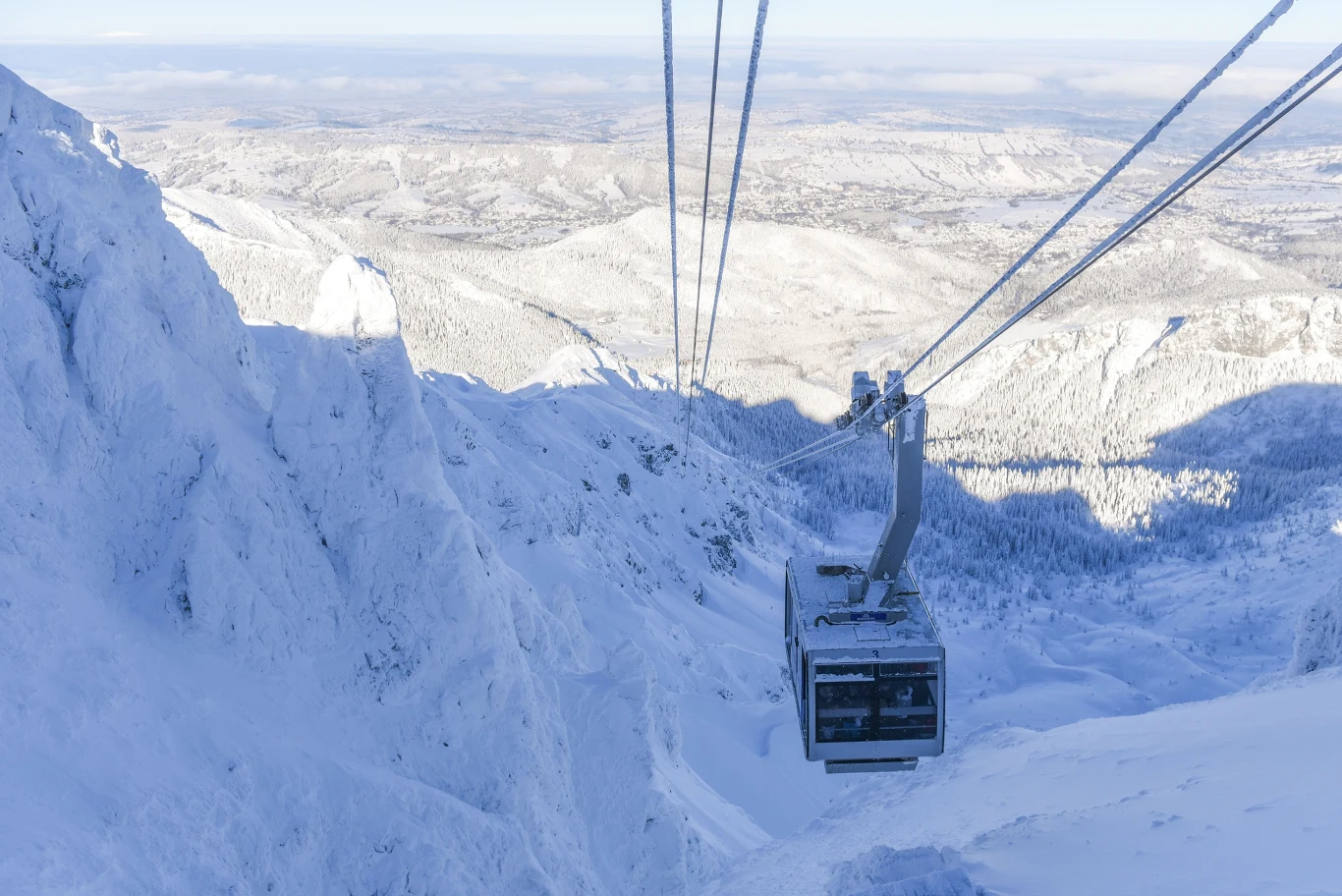 Kolejka linowa unosząca się nad ośnieżonym, górskim zboczem, otoczona jasnym, zimowym krajobrazem rozciągającym się po horyzont.